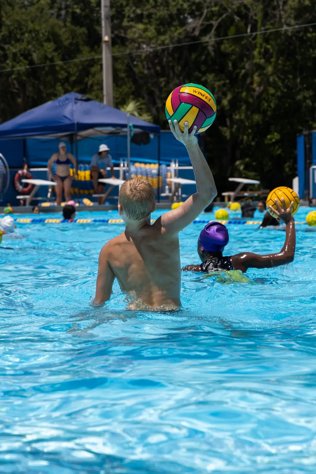 A camper raising the ball in the outdoor pool, tents and pines behind