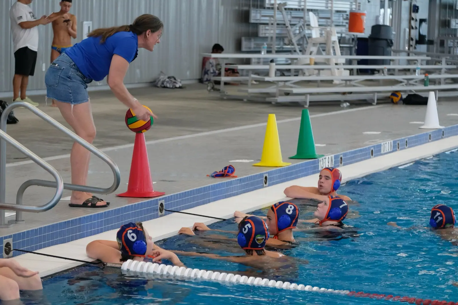 A coach setting out a ball for young players lined up along the pool wall