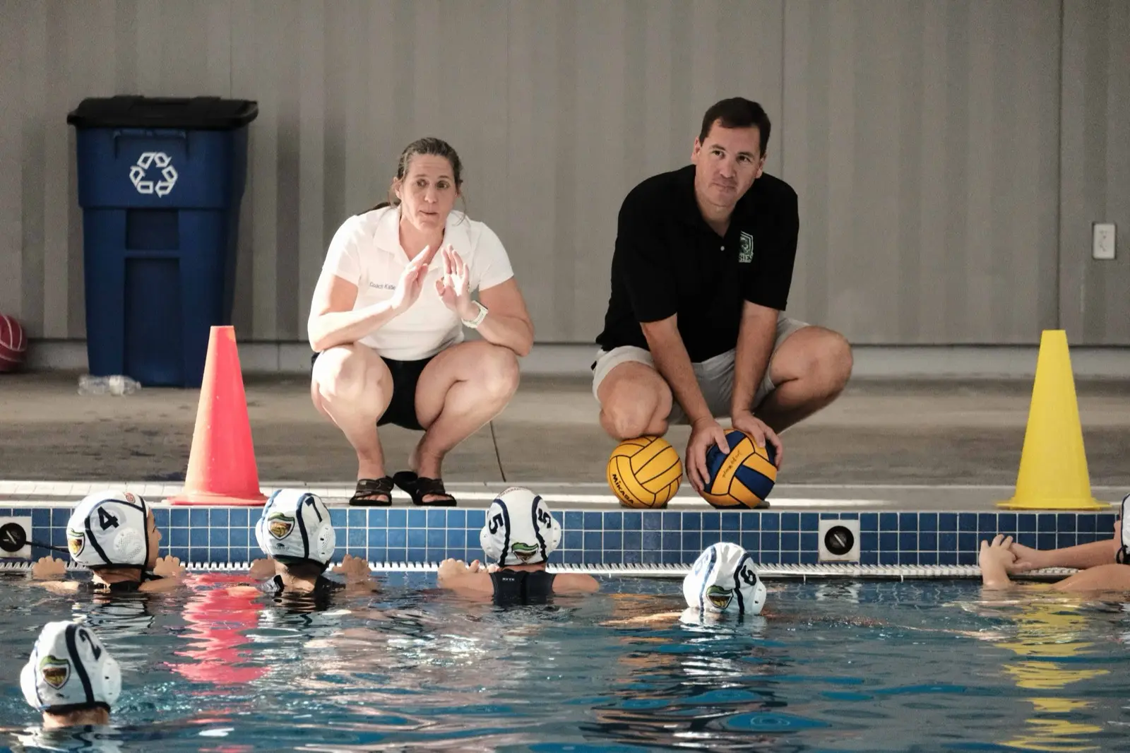 Two coaches crouched on deck briefing young players at the pool wall