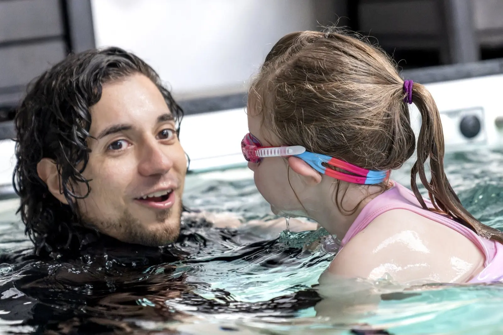 An instructor in the water listening closely to a young swimmer in pink goggles