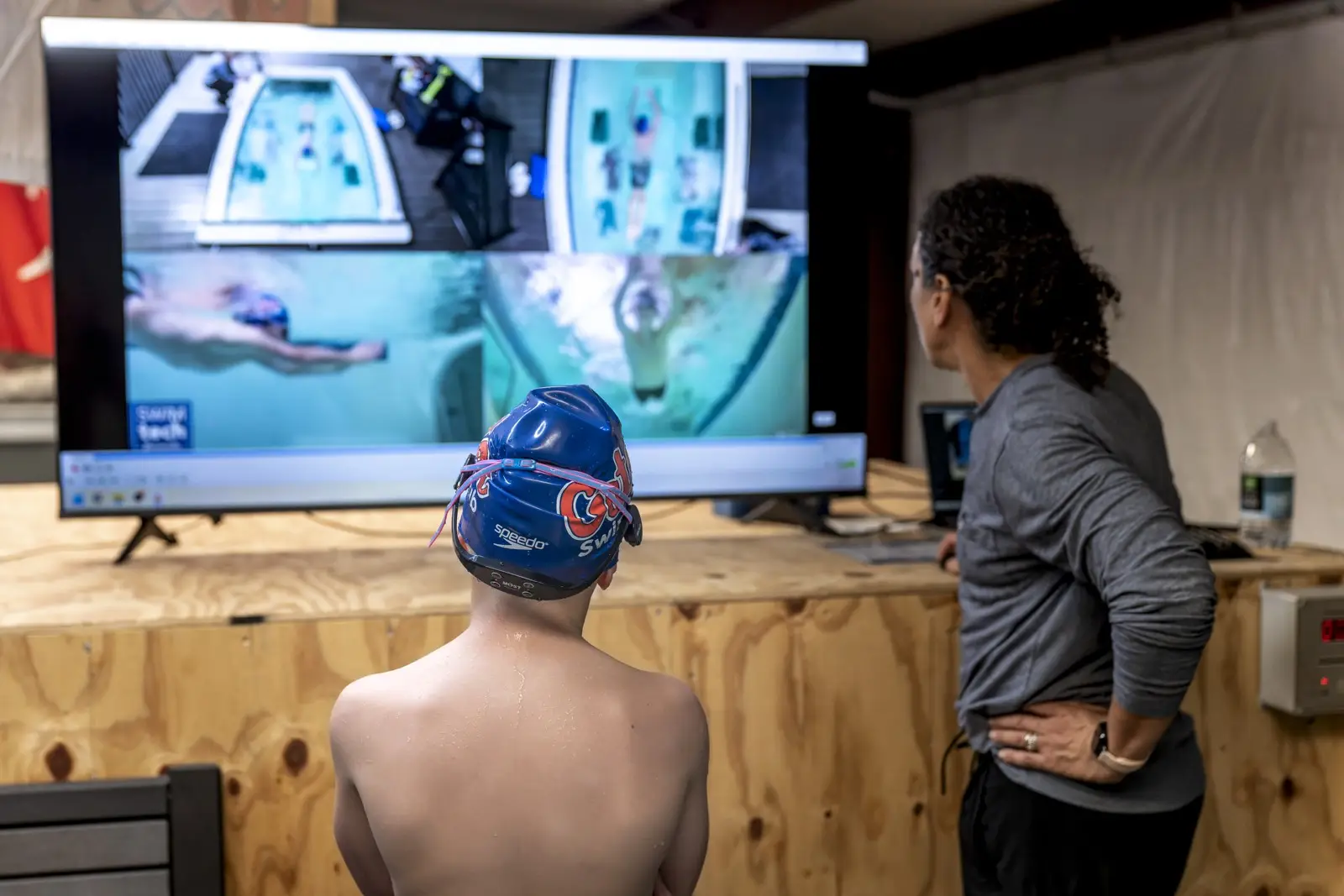 A coach and a teenage swimmer reviewing his stroke on a screen beside the pool