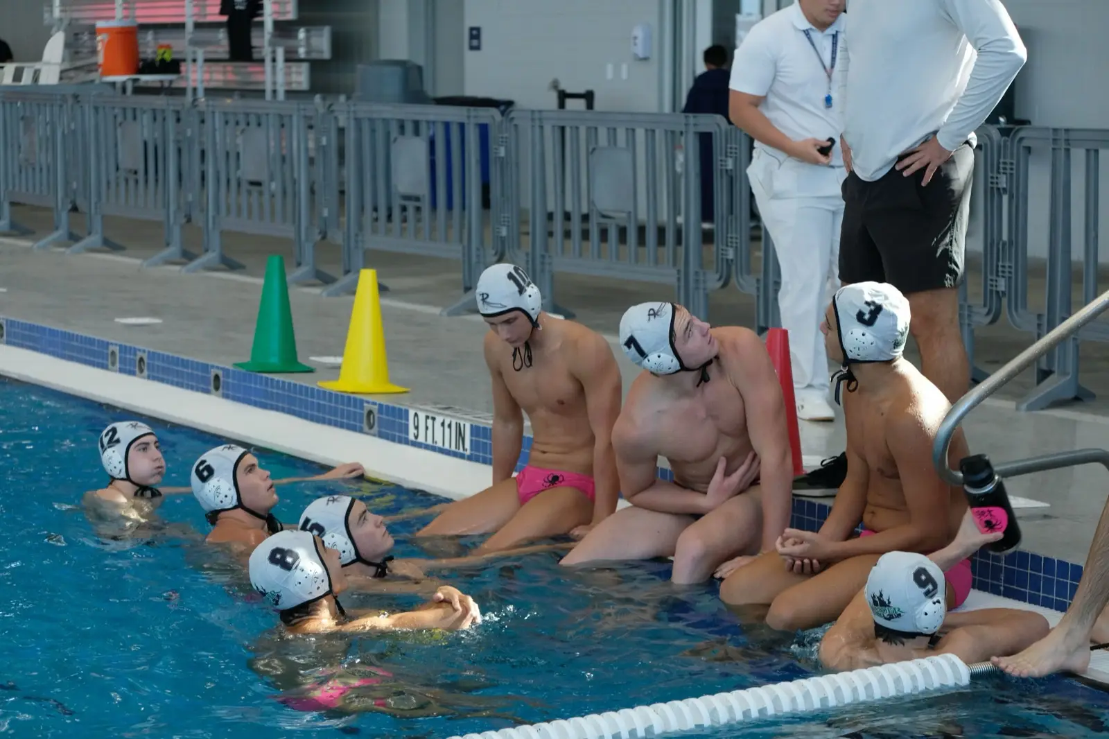 Young athletes in white caps lined up on the pool wall listening to their coach